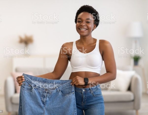 Slimming Motivation Concept. Skinny Black Female Showing Old Large Jeans After Successful Diet And Weight Loss Posing Smiling To Camera At Home. Weight-Loss Before And After Comparison